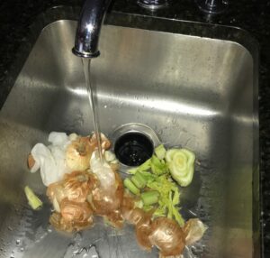 A stainless steel sink drain with the faucet on with onions and celery on the One Stop Plumbing garbage disposal page