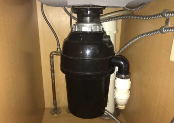 A White Garbage Disposal Under A Kitchen Cabinet With Black Pipes With A Link To One Stop Plumbing’s Garbage Disposal